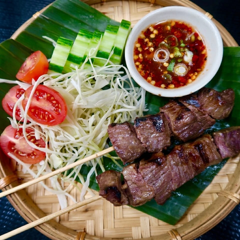 Grilled beef skewers with a spicy dipping sauce, shredded cabbage, cucumber, and tomato on a banana leaf.