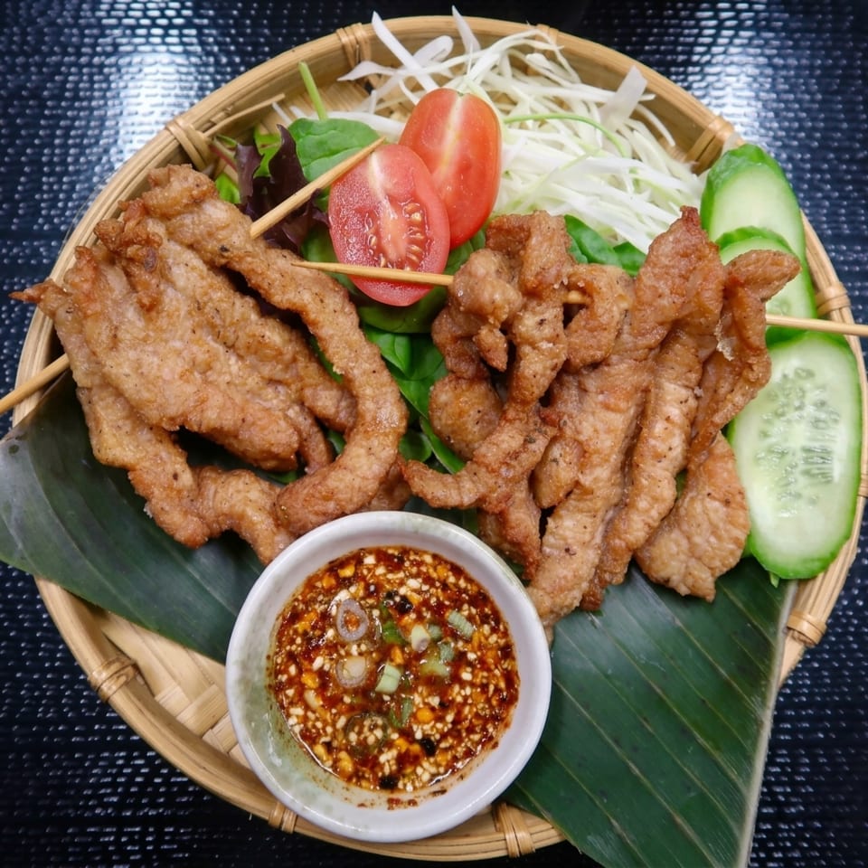 Fried pork skewers with dipping sauce, sliced tomatoes, cucumbers, and shredded cabbage on a bamboo tray.