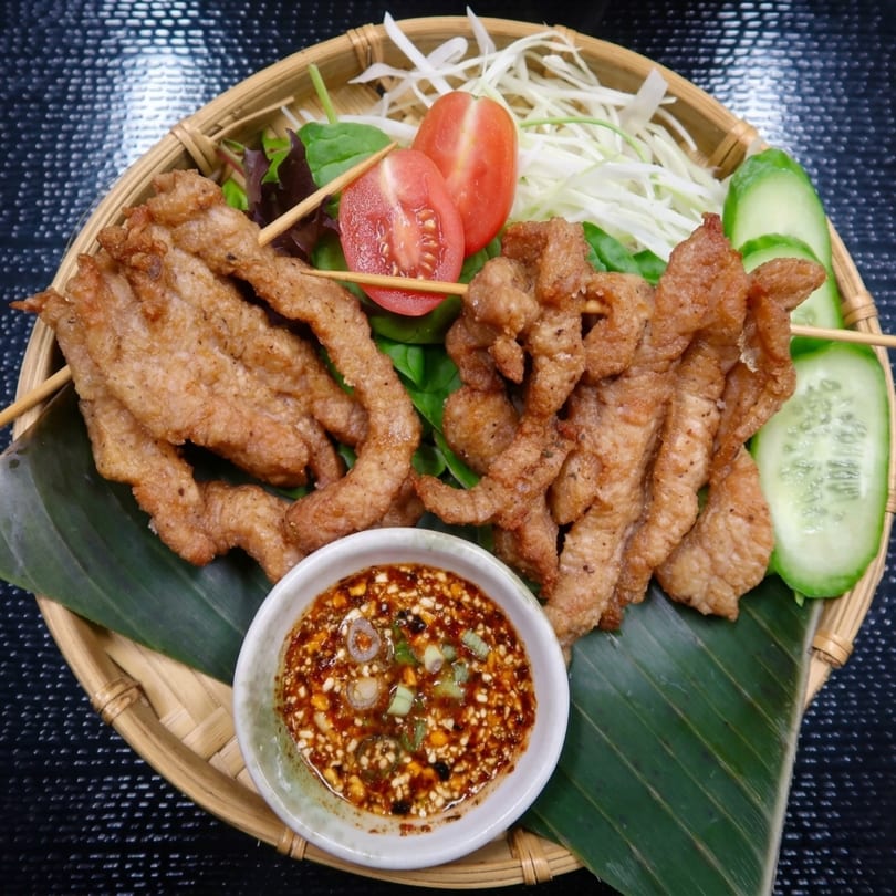 Fried pork skewers with dipping sauce, sliced tomatoes, cucumbers, and shredded cabbage on a bamboo tray.