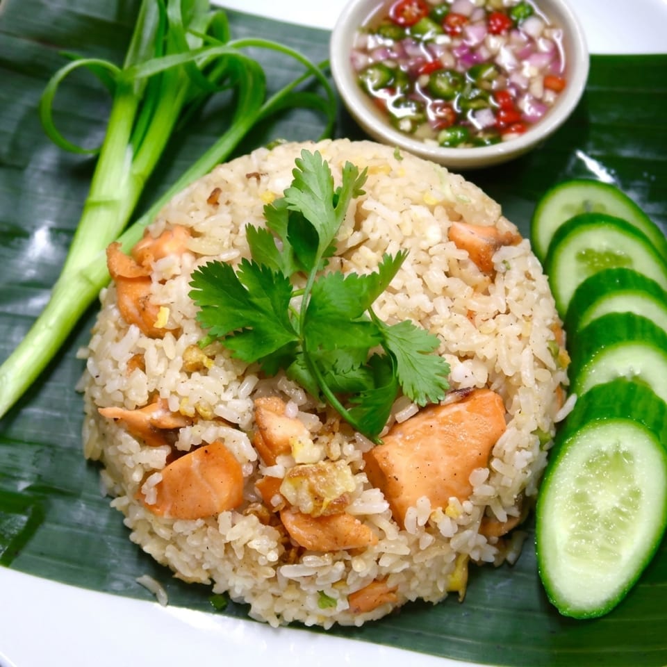 Salmon fried rice with cilantro, green onions, cucumber slices, and chili sauce on a banana leaf.